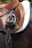 A close-up view of a brown leather horse saddle, featuring detailed stitching and attached metallic stirrups. The saddle sits atop a quilted white saddle pad placed on a dark-colored horse.