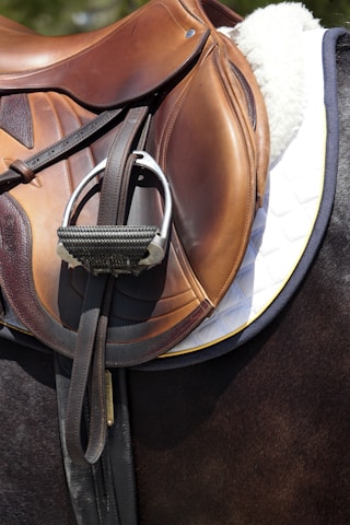 A close-up view of a brown leather horse saddle, featuring detailed stitching and attached metallic stirrups. The saddle sits atop a quilted white saddle pad placed on a dark-colored horse.