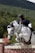 A person, dressed in a riding helmet and jacket, is engaged in equestrian jumping. They are mounted on a white horse that is leaping over a brick-patterned barrier. The background features a lush, green forest and a mountain under a partly cloudy sky.