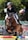 A rider in formal equestrian attire jumps over a colorful obstacle on a brown horse during a competition. There is a crowd of spectators in the background, with flags prominently displayed, indicating the event takes place outdoors.