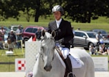 A person wearing a helmet and formal riding attire is mounted on a white horse. The scene appears to be part of an equestrian event, with spectators in the background and parked vehicles nearby. The outdoor setting includes grassy areas and a sunny climate.
