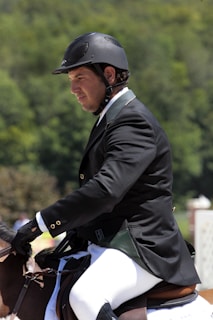 A person wearing a black riding helmet and a formal jacket sits on a horse, engaged in an equestrian activity. The background is a blurred mix of green foliage, suggesting an outdoor setting.
