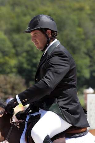 A person wearing a black riding helmet and a formal jacket sits on a horse, engaged in an equestrian activity. The background is a blurred mix of green foliage, suggesting an outdoor setting.