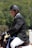 A person wearing a black riding helmet and a formal jacket sits on a horse, engaged in an equestrian activity. The background is a blurred mix of green foliage, suggesting an outdoor setting.
