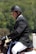 A person wearing a black riding helmet and a formal jacket sits on a horse, engaged in an equestrian activity. The background is a blurred mix of green foliage, suggesting an outdoor setting.