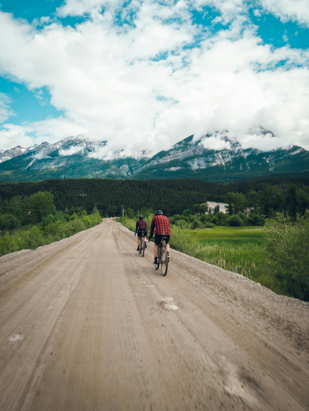 a couple of people riding bikes on a dirt road with mountains in the background