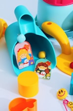 Brightly colored children's toys are scattered on a flat surface. There is a blue and yellow watering can, a yellow cylindrical container, a smiley face keychain, and two cards with illustrations of cartoon girls. Additional toys include a yellow ball, a cookie cutter, and a red ball.