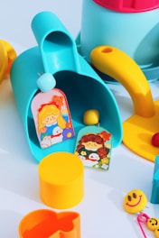 Brightly colored children's toys are scattered on a flat surface. There is a blue and yellow watering can, a yellow cylindrical container, a smiley face keychain, and two cards with illustrations of cartoon girls. Additional toys include a yellow ball, a cookie cutter, and a red ball.