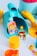 Brightly colored children's toys are scattered on a flat surface. There is a blue and yellow watering can, a yellow cylindrical container, a smiley face keychain, and two cards with illustrations of cartoon girls. Additional toys include a yellow ball, a cookie cutter, and a red ball.