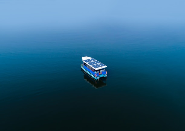 A small boat with solar panels on its roof floats on a vast, calm body of water. The scene is tranquil with a minimalistic composition and a clear horizon.