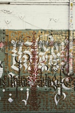 Elaborate graffiti art featuring intricate calligraphy is displayed on a brick wall. The design includes layered text in various styles and colors, such as black, red, and gold. The wall has a distressed and urban aesthetic, with a mix of geometric shapes and patterns. There is also a large vertical pipe or pole on the right side, covered with numerous colorful stickers.