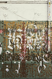 Elaborate graffiti art featuring intricate calligraphy is displayed on a brick wall. The design includes layered text in various styles and colors, such as black, red, and gold. The wall has a distressed and urban aesthetic, with a mix of geometric shapes and patterns. There is also a large vertical pipe or pole on the right side, covered with numerous colorful stickers.