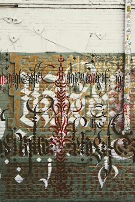 Elaborate graffiti art featuring intricate calligraphy is displayed on a brick wall. The design includes layered text in various styles and colors, such as black, red, and gold. The wall has a distressed and urban aesthetic, with a mix of geometric shapes and patterns. There is also a large vertical pipe or pole on the right side, covered with numerous colorful stickers.