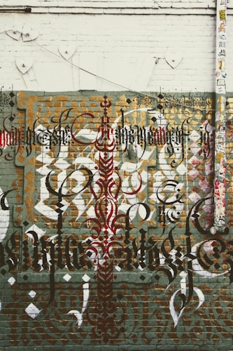 Elaborate graffiti art featuring intricate calligraphy is displayed on a brick wall. The design includes layered text in various styles and colors, such as black, red, and gold. The wall has a distressed and urban aesthetic, with a mix of geometric shapes and patterns. There is also a large vertical pipe or pole on the right side, covered with numerous colorful stickers.