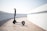 A modern electric scooter is parked on a wooden boardwalk, leaning slightly to one side. The background features a clean and minimalistic white wall curving around the path.