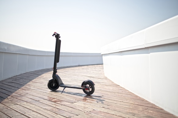 A modern electric scooter is parked on a wooden boardwalk, leaning slightly to one side. The background features a clean and minimalistic white wall curving around the path.