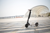 An electric scooter is parked on a paved area with a modern, white, curved architectural structure in the background. The scene is spacious with long shadows cast on the ground, suggesting a sunny day. A distant view of a bridge is also visible.