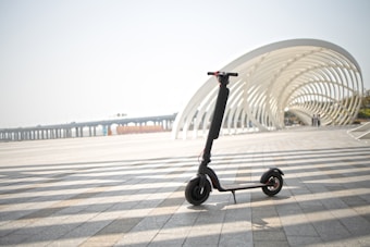 An electric scooter is parked on a paved area with a modern, white, curved architectural structure in the background. The scene is spacious with long shadows cast on the ground, suggesting a sunny day. A distant view of a bridge is also visible.