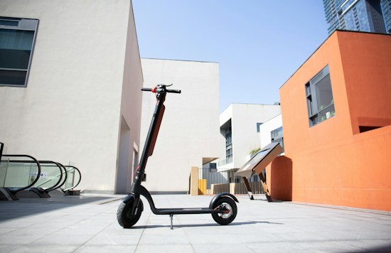 A black electric scooter is parked on a modern urban plaza with geometric buildings. The architecture features contrasting white and orange walls, with large windows and flat surfaces. The sky is clear and blue, suggesting a sunny day.