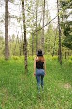 A person wearing a black sleeveless top, blue jeans, and a black hat walks through a lush forest. The environment is dense with green grass and tall trees, indicating a vibrant and healthy ecosystem. The person is seen from behind as they move through the natural landscape.