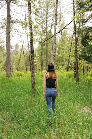 A person wearing a black sleeveless top, blue jeans, and a black hat walks through a lush forest. The environment is dense with green grass and tall trees, indicating a vibrant and healthy ecosystem. The person is seen from behind as they move through the natural landscape.