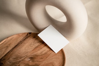 A minimalist composition featuring a wooden tray, a small white card, and a circular ceramic object with a hole in the center. The objects are placed on a neutral-toned fabric background.
