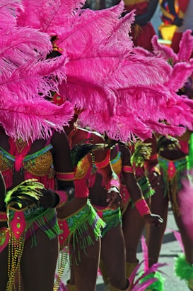 A group of dancers in vibrant, colorful costumes adorned with large pink feather headdresses and intricate beadwork. The costumes feature bright neon green and pink details with hints of gold. The dancers hold green and pink feathered accessories, adding to the lively and festive atmosphere. The scene likely depicts a carnival or parade setting, filled with movement and energy.