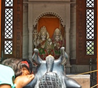 Two intricately decorated statues of Hindu deities are positioned within a shrine adorned with ornate carvings. A person is seen bowing in prayer towards a black statue of Nandi in the foreground.