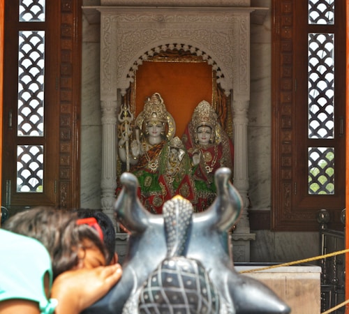 Two intricately decorated statues of Hindu deities are positioned within a shrine adorned with ornate carvings. A person is seen bowing in prayer towards a black statue of Nandi in the foreground.