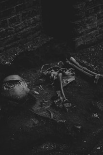 A dark and moody scene showing human skeletal remains on the ground against a brick wall. The bones appear scattered and partially buried in dirt, conveying an eerie and abandoned atmosphere.