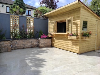 A wooden garden shed with open windows surrounded by a neatly landscaped area. The shed is made of light-colored wood and features flower boxes with vibrant blooms. Behind the shed, there is a high stone wall with lattice panels and various plants. The patio area in front is tiled with light-colored slabs, and the space is filled with sunlight.