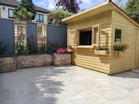 A wooden garden shed with open windows surrounded by a neatly landscaped area. The shed is made of light-colored wood and features flower boxes with vibrant blooms. Behind the shed, there is a high stone wall with lattice panels and various plants. The patio area in front is tiled with light-colored slabs, and the space is filled with sunlight.
