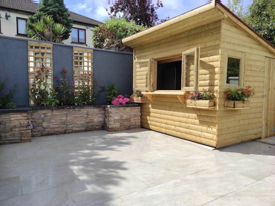 A wooden garden shed with open windows surrounded by a neatly landscaped area. The shed is made of light-colored wood and features flower boxes with vibrant blooms. Behind the shed, there is a high stone wall with lattice panels and various plants. The patio area in front is tiled with light-colored slabs, and the space is filled with sunlight.
