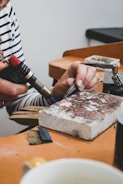 A person is seen working on a crafting or jewelry-making project. They are using a blowtorch and holding a metal tool to work on a piece of metal or jewelry placed on a brick. The workbench has various tools and materials scattered around, with a white cup partially visible in the foreground. The person is wearing a striped long-sleeve shirt.