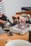 A person is seen working on a crafting or jewelry-making project. They are using a blowtorch and holding a metal tool to work on a piece of metal or jewelry placed on a brick. The workbench has various tools and materials scattered around, with a white cup partially visible in the foreground. The person is wearing a striped long-sleeve shirt.