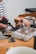 A person is seen working on a crafting or jewelry-making project. They are using a blowtorch and holding a metal tool to work on a piece of metal or jewelry placed on a brick. The workbench has various tools and materials scattered around, with a white cup partially visible in the foreground. The person is wearing a striped long-sleeve shirt.