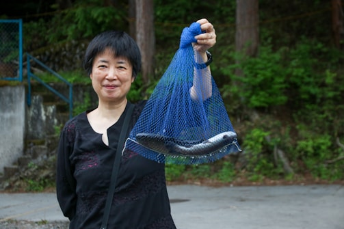 A person holding a blue net containing two fish, with a forested background. The person is smiling and is wearing a dark outfit.
