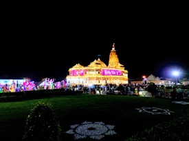 A brightly lit temple decorated with colorful lights during the night. A large crowd is gathered in front of the temple, and there are decorations on the ground.