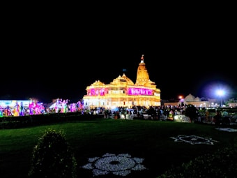 A brightly lit temple decorated with colorful lights during the night. A large crowd is gathered in front of the temple, and there are decorations on the ground.