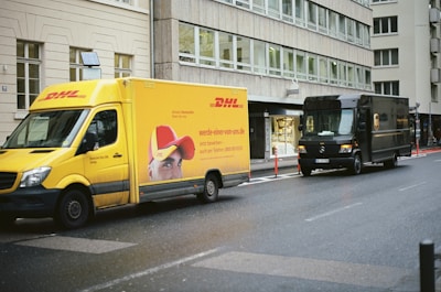 Two delivery trucks are parked on a city street. The truck on the left is yellow with DHL branding, displaying a partial image of a person wearing a red cap. The truck on the right is dark brown with UPS branding. The street is wet, suggesting recent rain, and the setting appears to be urban, with multi-story buildings lining the street.