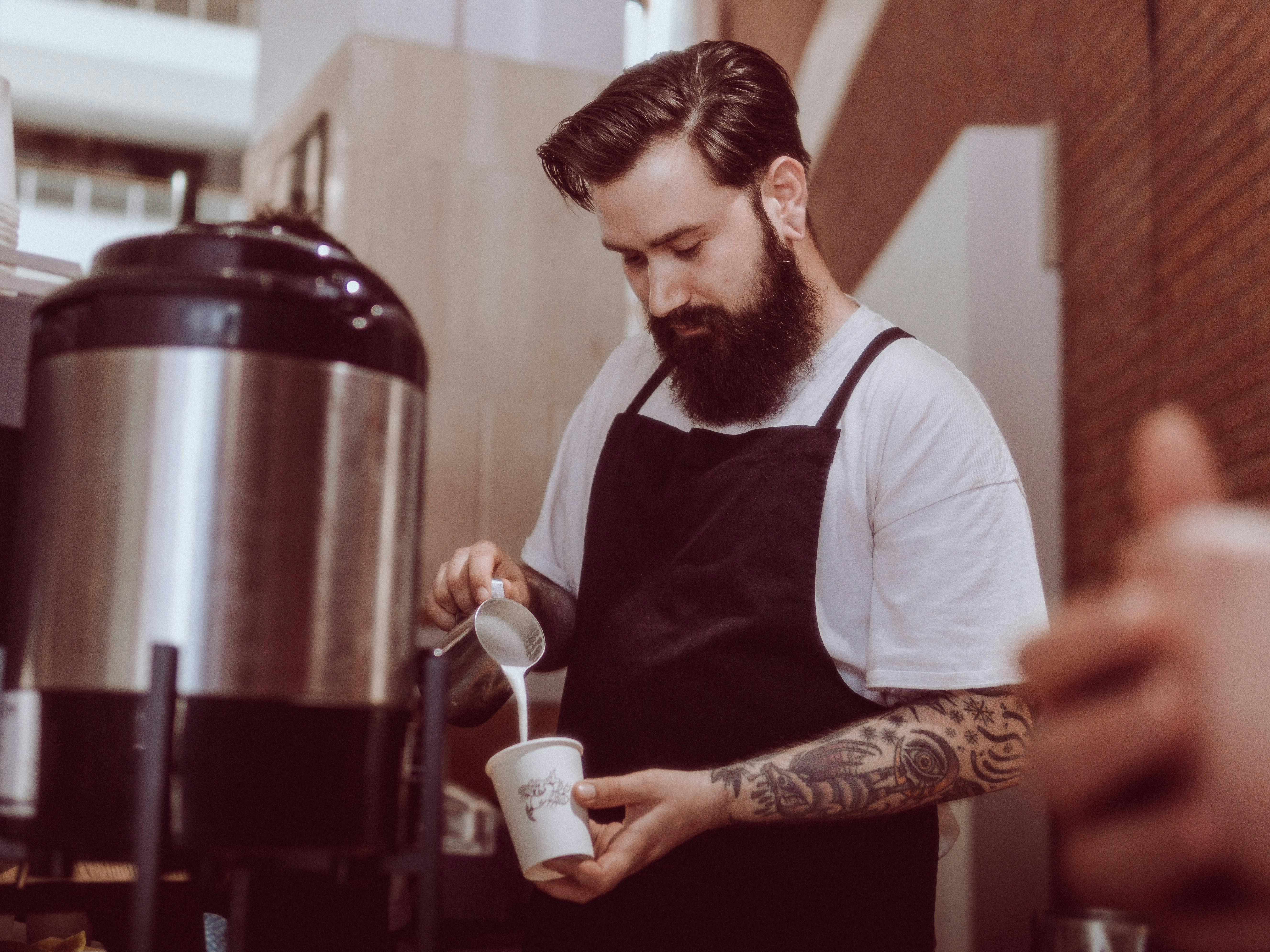 Bearded barista in apron steaming milk beside an urn