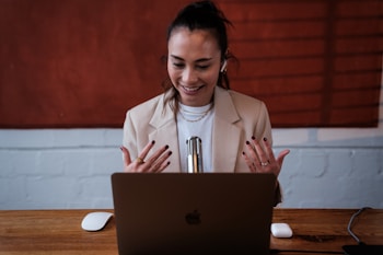 A woman is seated at a desk, engaged in a video call or recording session using a laptop. She is smiling and gesturing with her hands, wearing a beige blazer, and there is a microphone placed in front of her. The background features a textured red décor and a white brick wall.