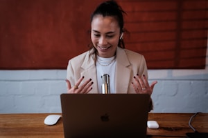 A woman is seated at a desk, engaged in a video call or recording session using a laptop. She is smiling and gesturing with her hands, wearing a beige blazer, and there is a microphone placed in front of her. The background features a textured red d&eacute;cor and a white brick wall.