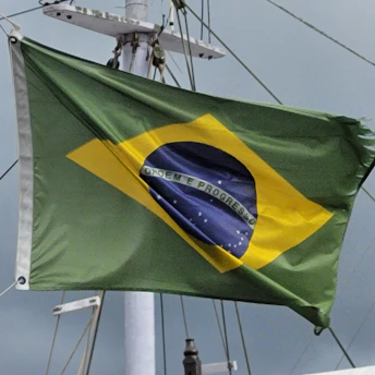 A flag with a green field, a yellow diamond at the center, and a blue globe with white stars depicted inside. The globe has a white banner across it, with the words 'Ordem e Progresso' written on it. The flag is attached to a pole with visible rigging and is seen against a cloudy sky.