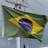 A flag with a green field, a yellow diamond at the center, and a blue globe with white stars depicted inside. The globe has a white banner across it, with the words 'Ordem e Progresso' written on it. The flag is attached to a pole with visible rigging and is seen against a cloudy sky.