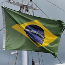 A flag with a green field, a yellow diamond at the center, and a blue globe with white stars depicted inside. The globe has a white banner across it, with the words 'Ordem e Progresso' written on it. The flag is attached to a pole with visible rigging and is seen against a cloudy sky.