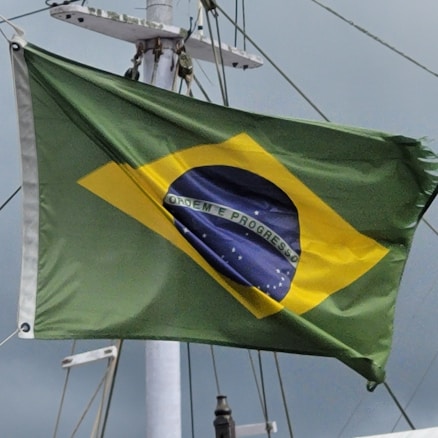 A flag with a green field, a yellow diamond at the center, and a blue globe with white stars depicted inside. The globe has a white banner across it, with the words 'Ordem e Progresso' written on it. The flag is attached to a pole with visible rigging and is seen against a cloudy sky.