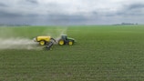 A green tractor is towing a large yellow agricultural sprayer across a vast field of crops under an overcast sky. Dust is visible behind the equipment, indicating movement and activity in tending to the field.