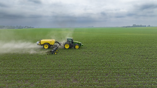 A green tractor is towing a large yellow agricultural sprayer across a vast field of crops under an overcast sky. Dust is visible behind the equipment, indicating movement and activity in tending to the field.