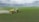 A green tractor is towing a large yellow agricultural sprayer across a vast field of crops under an overcast sky. Dust is visible behind the equipment, indicating movement and activity in tending to the field.